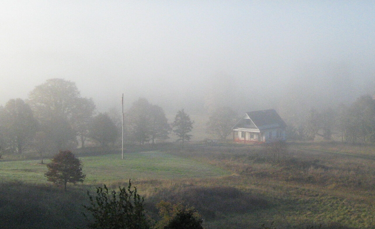 A house sits on a foggy morning surrounded by a field and forest, with mountains in the background