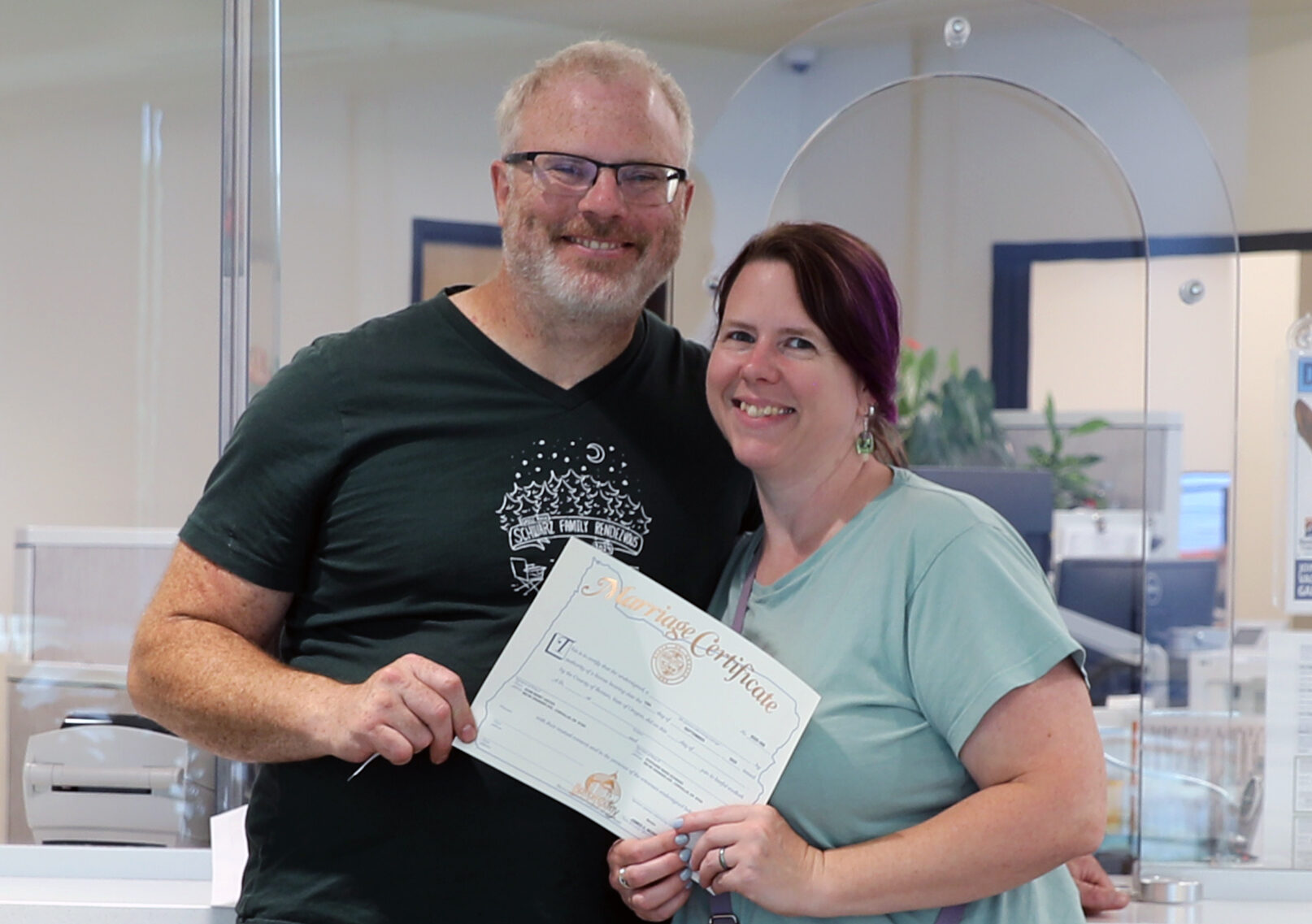 A man and a woman standing together holding a marriage certificate in front of the county's records services counter