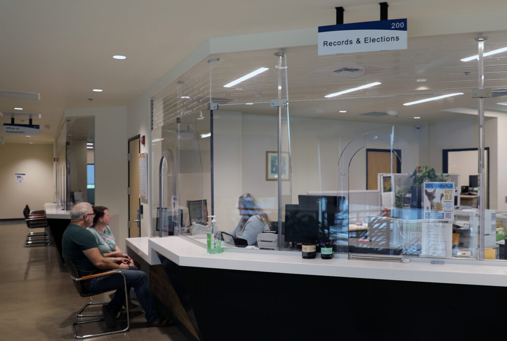 A couple sits at a sleek counter being helped by a Records & Elections employee behind the counter. Above the area on the ceiling hangs a sign that reads "Records & Elections. 200."