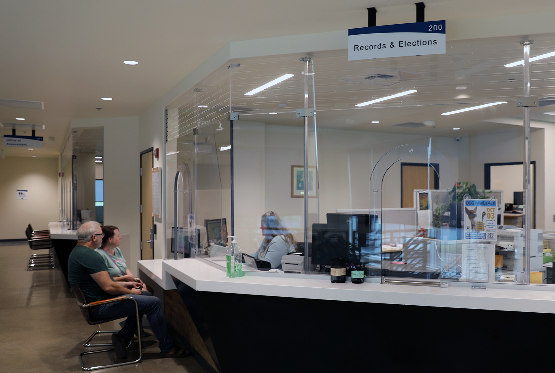 A couple sits at a sleek counter being helped by a Records & Elections employee behind the counter. Above the area on the ceiling hangs a sign that reads "Records & Elections. 200."