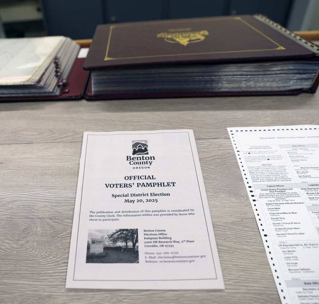 A piece of paper on a desk, in front of a large Records & Elections book. The Paper is the front page of Benton County's Official Voter Pamphlet for the General Elections Nov 5th, 2024. Text reads "The publication and distribution of this pamphlet is coordinated by the County Clerk. The information within was provided by those who chose to participate."