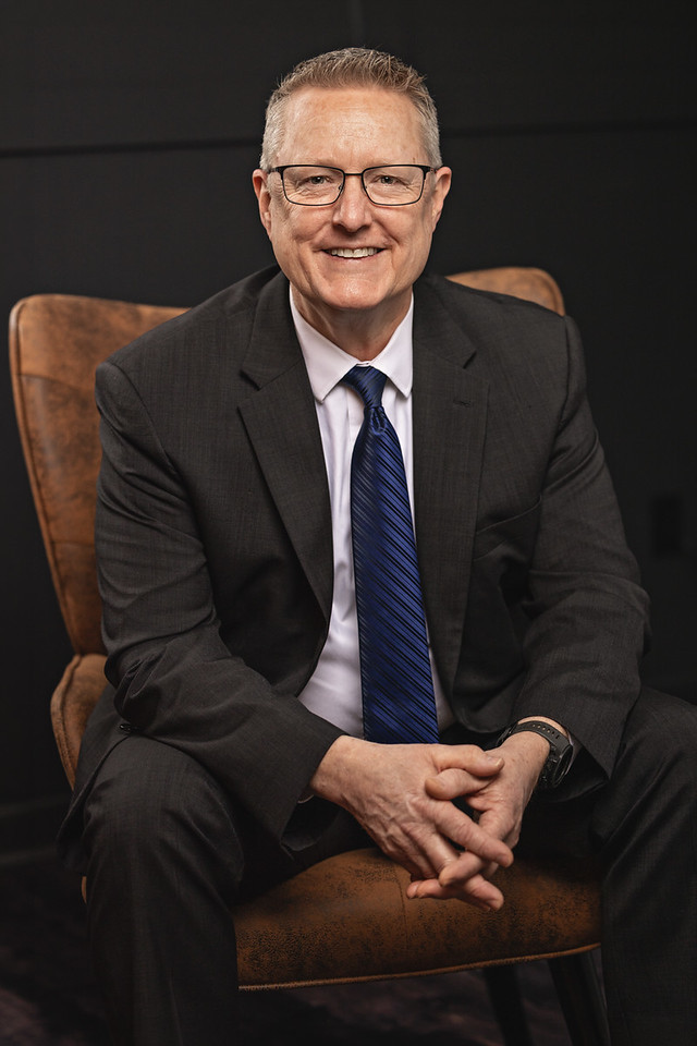 Man in a suit with a blue tie sits on a leather chair, smiling kindly at viewer