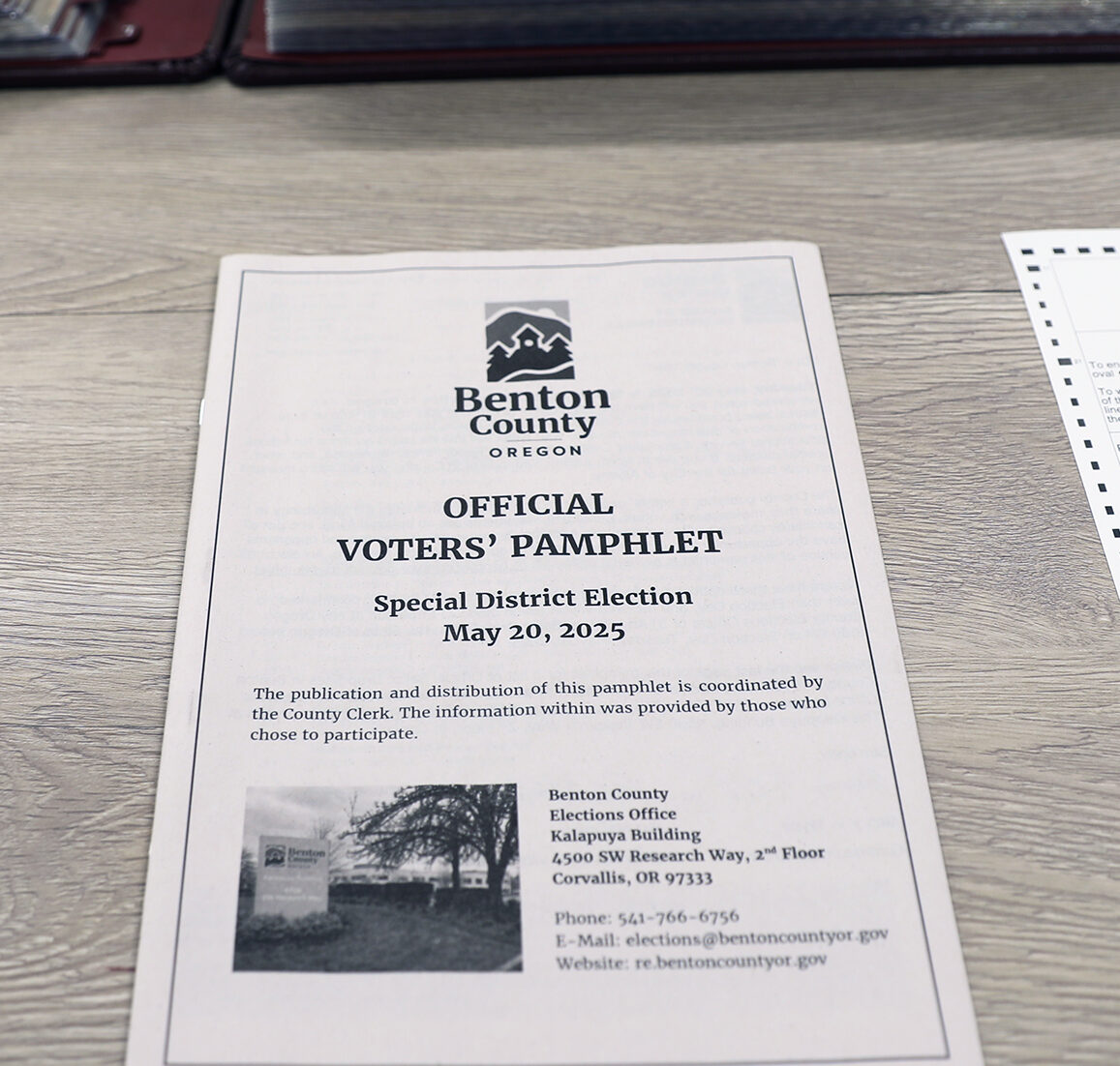 A piece of paper on a desk, in front of a large Records & Elections book. The Paper is the front page of Benton County's Official Voter Pamphlet for the General Elections Nov 5th, 2024. Text reads "The publication and distribution of this pamphlet is coordinated by the County Clerk. The information within was provided by those who chose to participate."